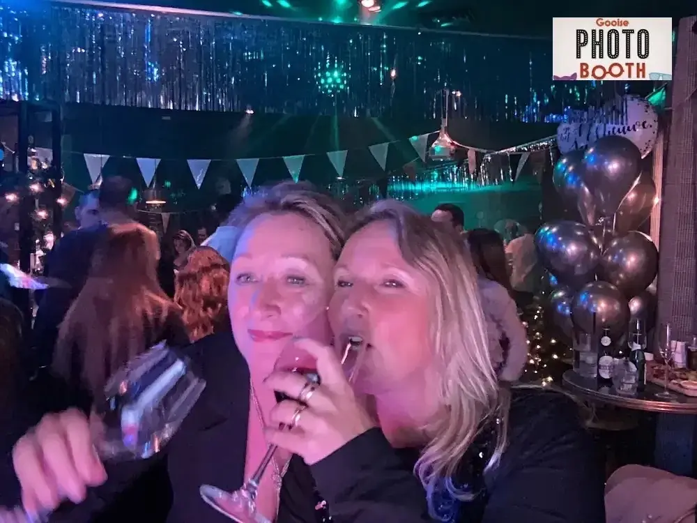 Twee vrouwen met een glas op een feest, foto gemaakt met de Gooise Photobooth, met ballonnen en feestverlichting op de achtergrond